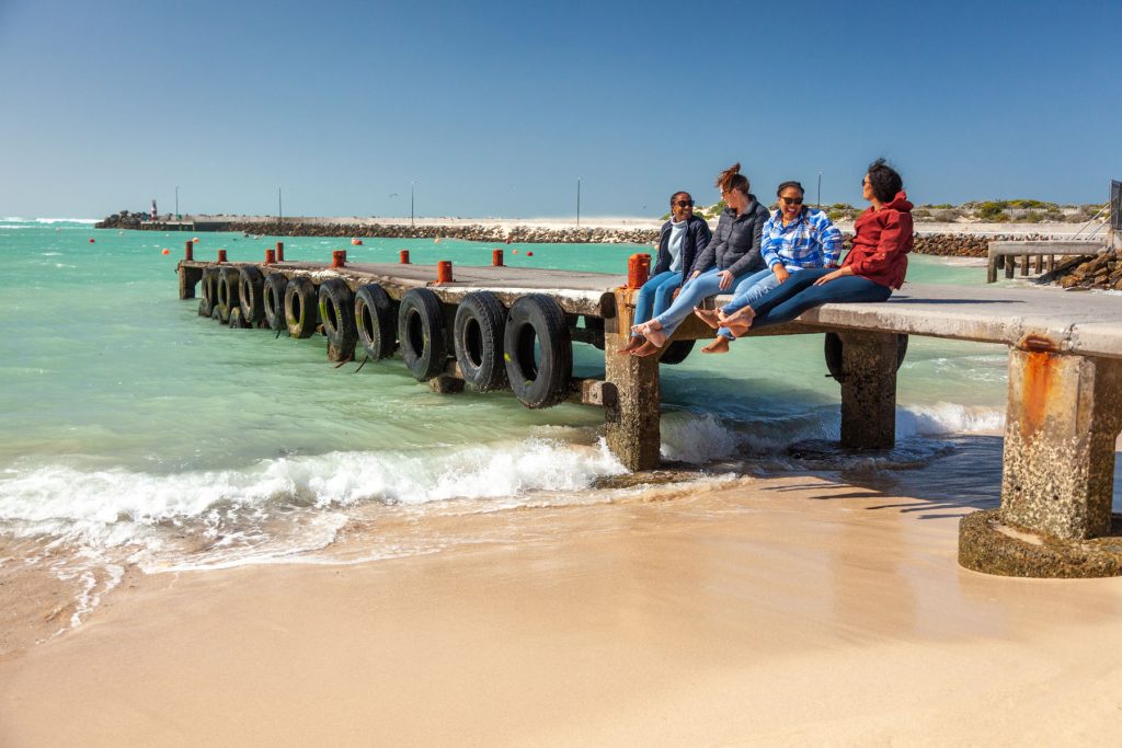 Four people sitting on the struisbaai harbour pier promoting where to stay in Cape Agulhas.
