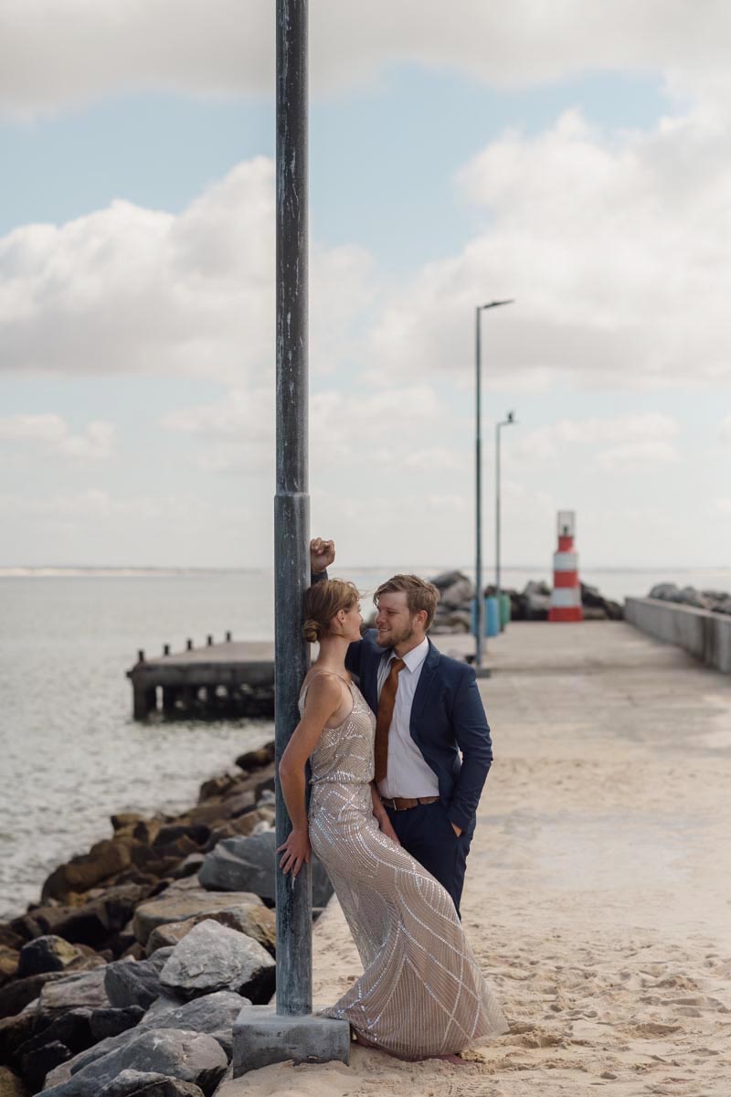 couple taking a wedding photos on the struisbaai harbour dock, with a lighthouse in the background.
